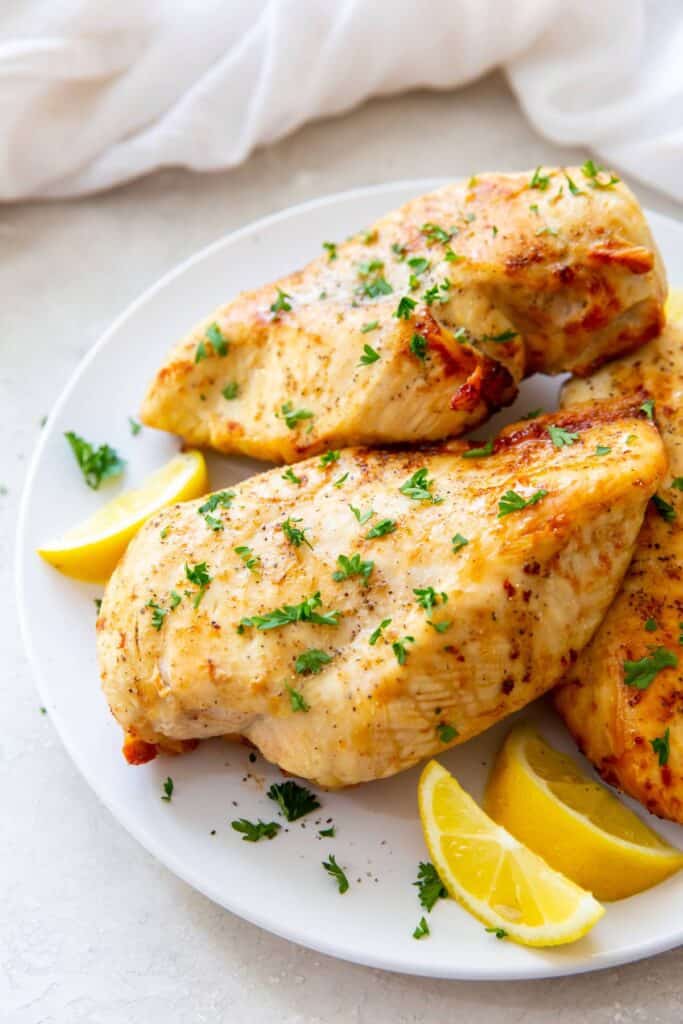 Unsliced air fry chicken breasts on a white plate with lemon wedges.