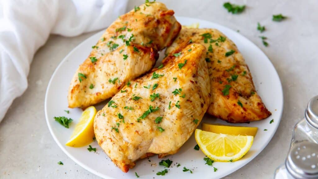 Cooked air fried chicken breast on a plate topped with parsley and next to lemon wedges.