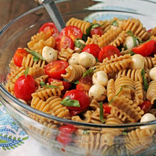 Caprese pasta salad in a clear bowl with serving spoon inside it to serve cold for lunch.