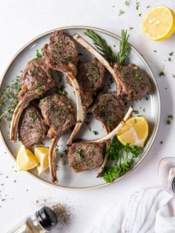 Overview shot of lamb chops served together on a white plate with lemon wedges and green garnishes