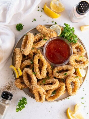 Zoomed out view of the breaded calamari on a serving platter with cocktail sauce and lemon wedges and green garnish around it