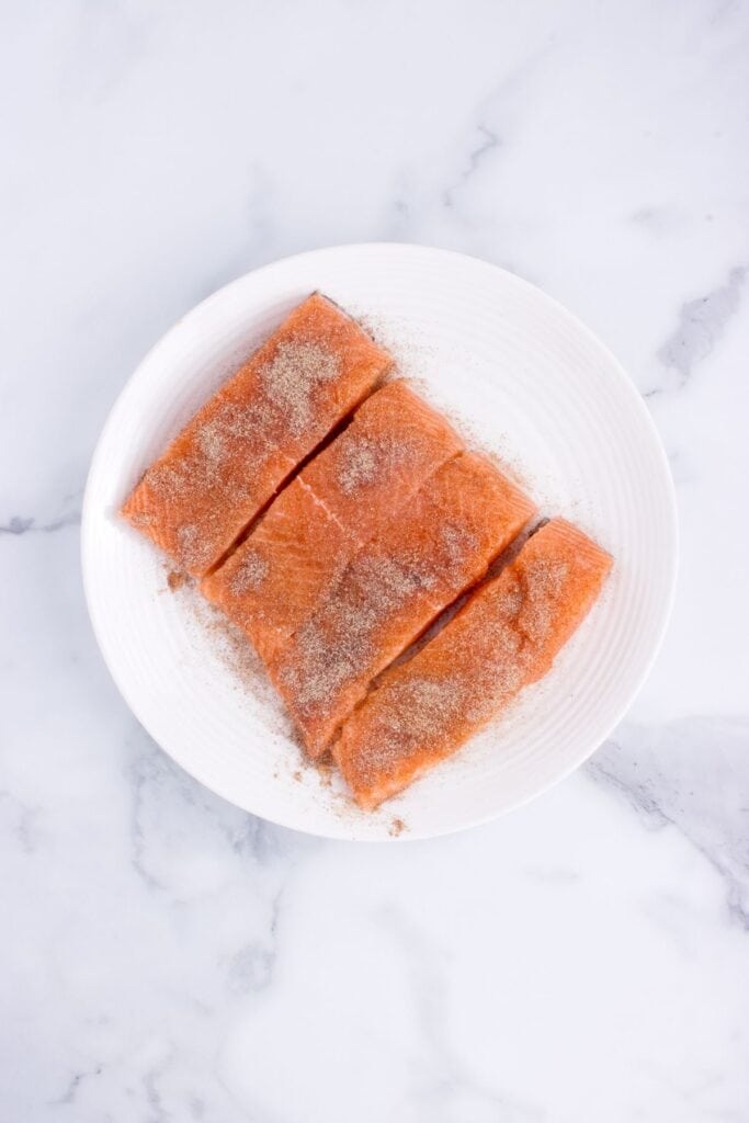 Salmon being seasoned on a white plate.