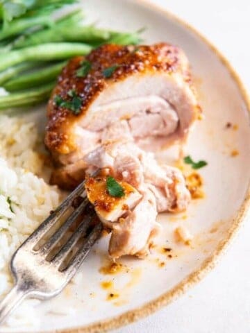 A fork holding a piece of boneless chicken thigh on a white plate with green beans in the background.