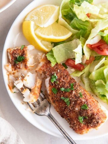 Cooked cod on a white plate with pieces flaked. Small salad and lemon wedges in the background