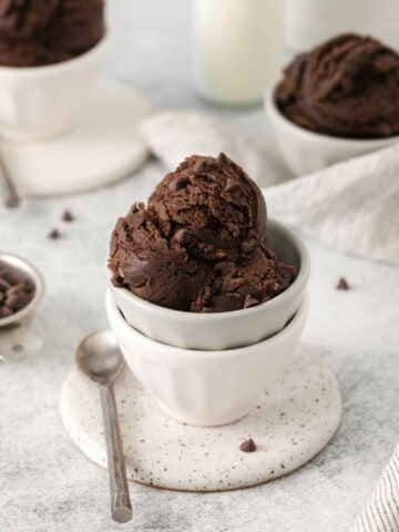 Scoops of edible brownie batter in a white bowl on a plate with a spoon resting on the plate.