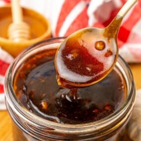 A spoon lifting a small amount of Honey Garlic Sauce out of a small clear mason jar.