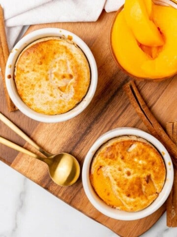 Two peach cobblers in white ramekins resting on a wooden board with peaches in the background.