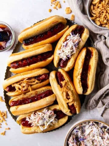 Overhead view of eight grilled bbq hot dogs on a plate with buns and toppings.