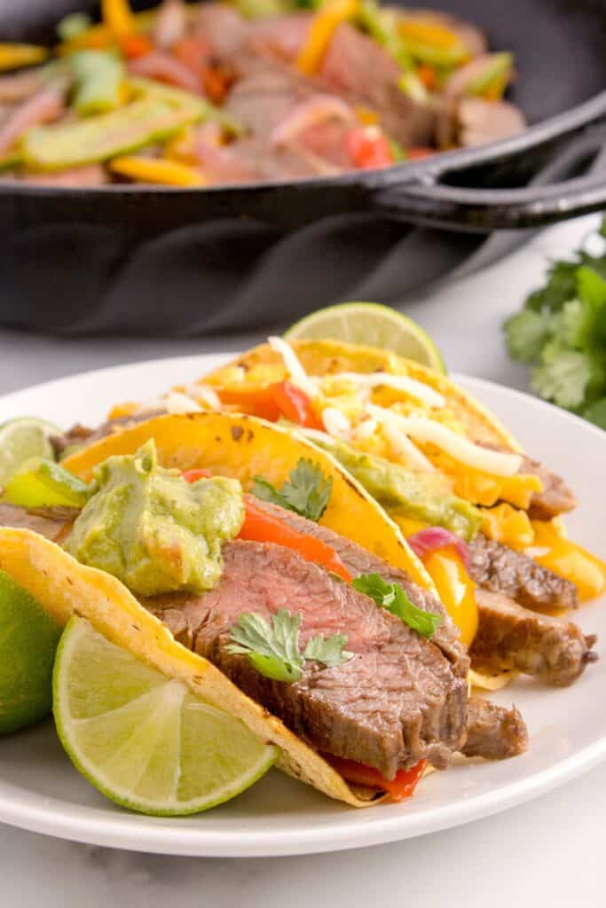 Steak fajitas in corn tortillas topped with a dollop of guacamole and a lime wedge.