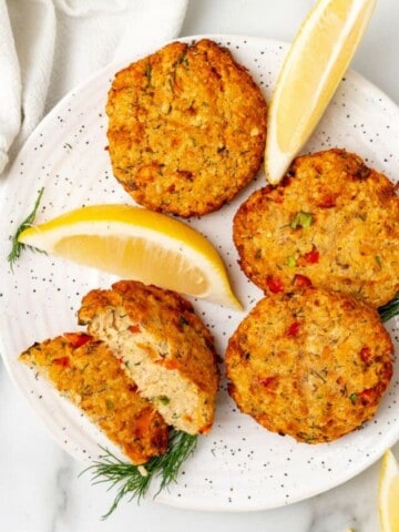 Three whole salmon patties, one salmon patty sliced in half on a white plate with lemon slices.