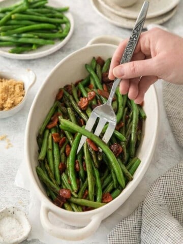 A fork lifting a bite of green beans with bacon from an oblong baking dish.