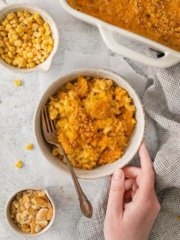 A bowl of macaroni corn casserole surrounded by cheese and the casserole dish.