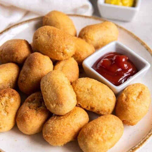 Miniature corn dogs on a white plate with ketchup and mustard for dipping.