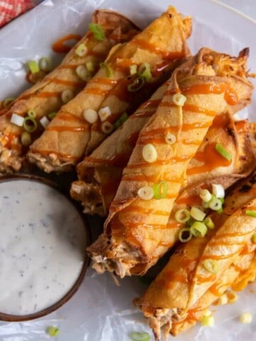 A stack of prepared taquitos topped with green onions and drizzled with buffalo sauce.