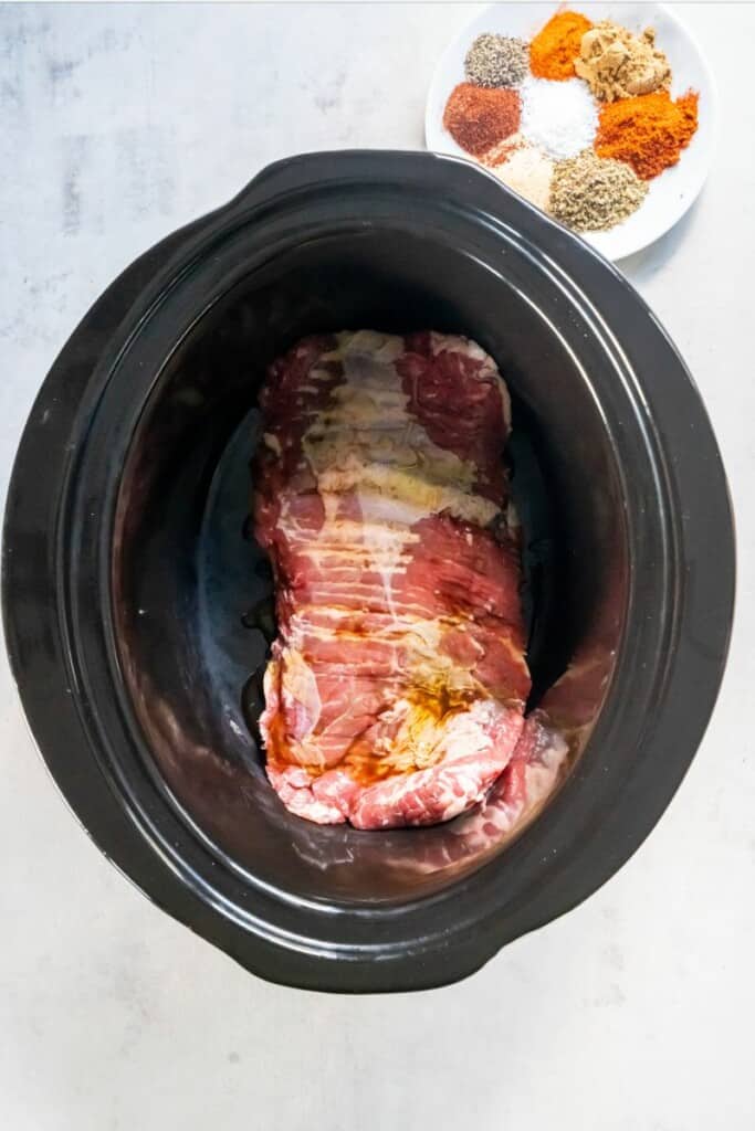Skirt steak in a black crock pot with a small white plate holding the seasonings next to it.