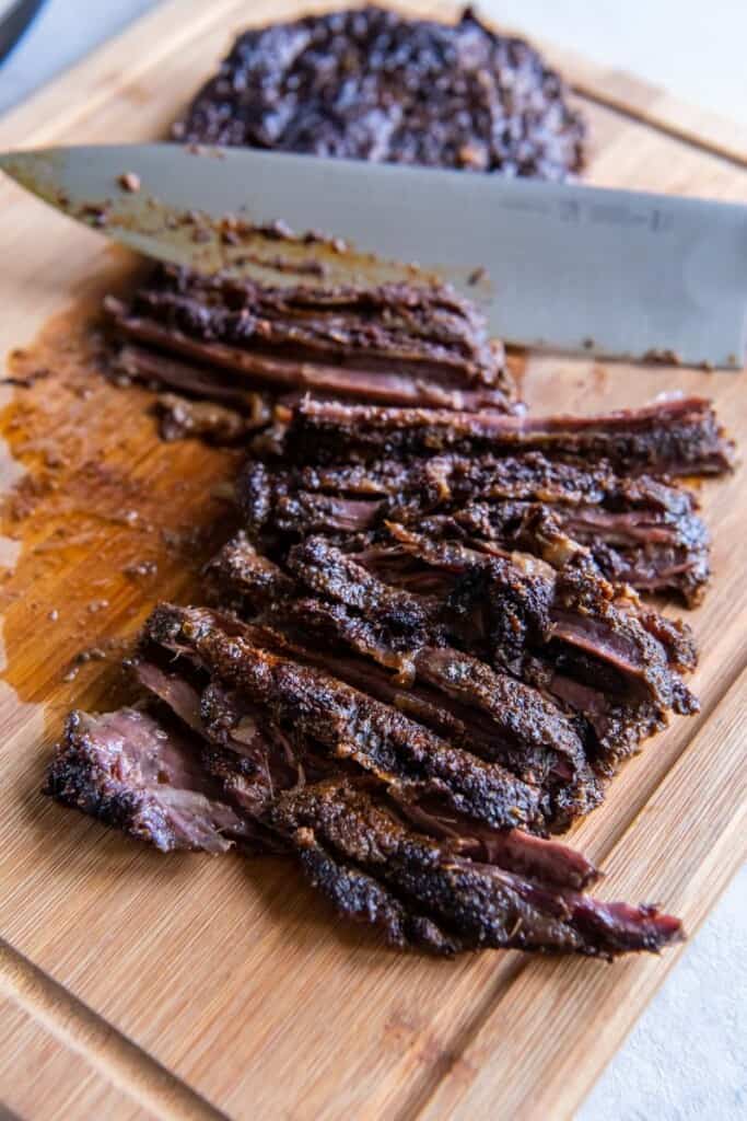 A chef's knife cutting a skirt steak into slices on a wooden cutting board.