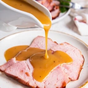 pouring ham gravy over slices of ham on a plate.