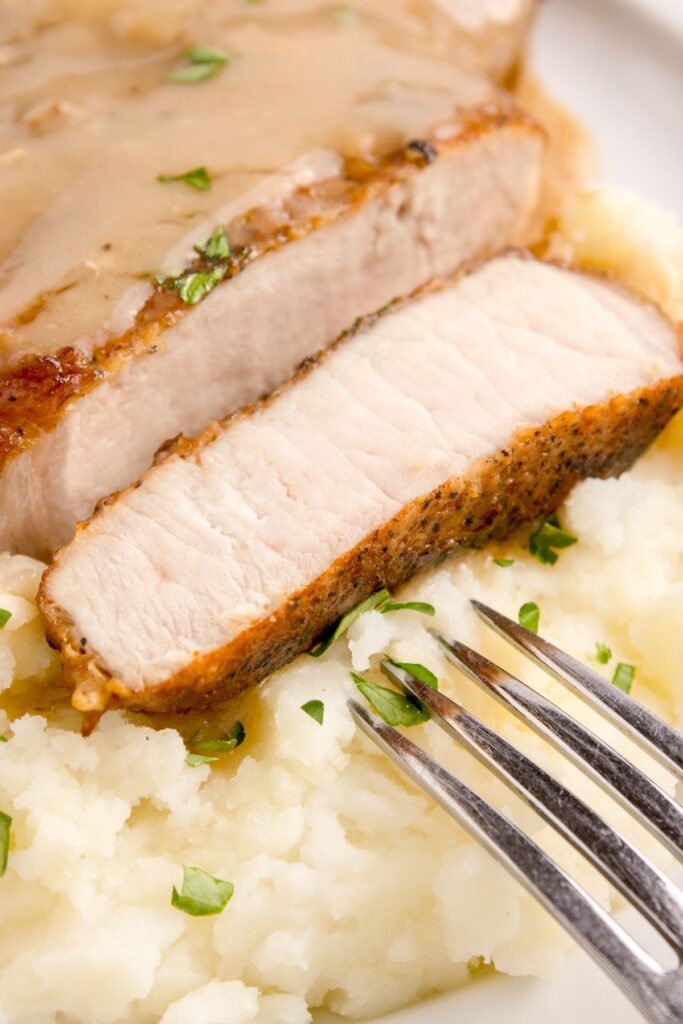 Smothered pork chop cut to see the inside sitting on top of mashed potatoes with a fork.