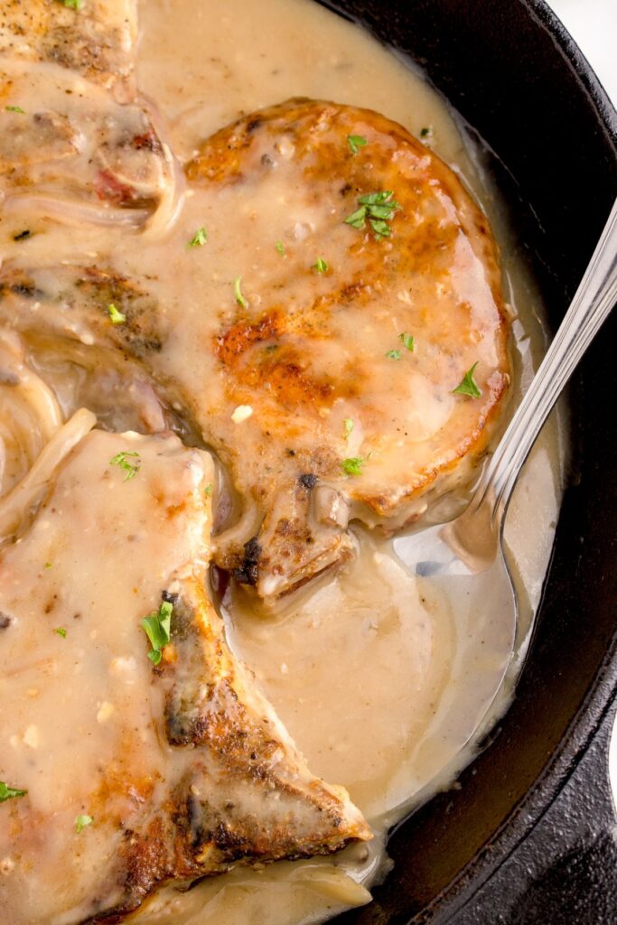 Closeup of the smothered pork chops in a cast iron skillet with the gravy on top with a spoon in the pan.