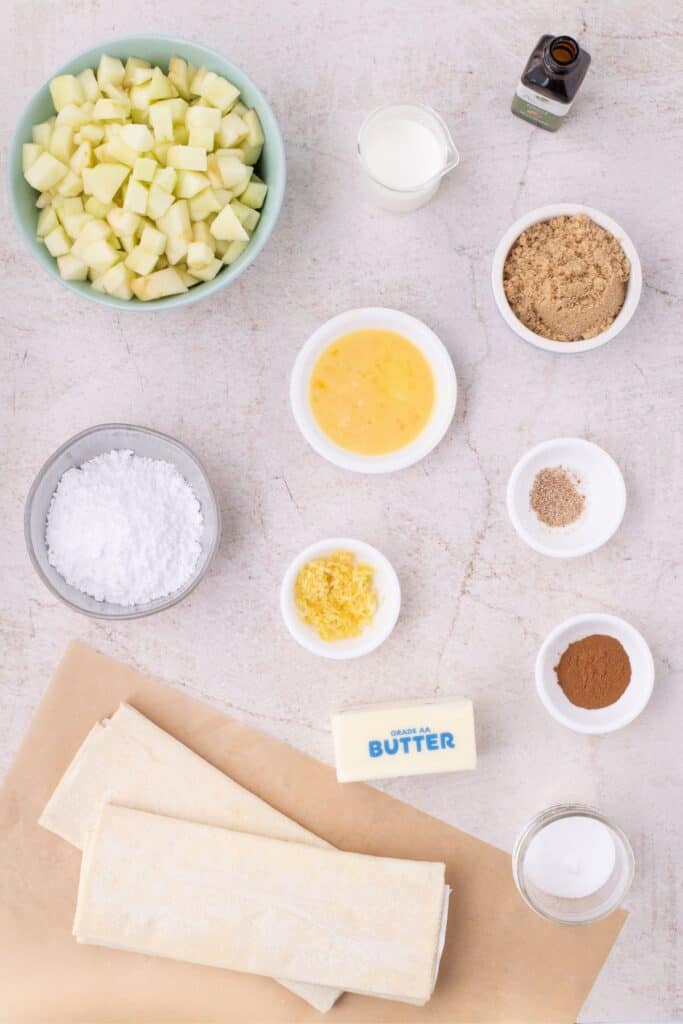 Flatlay with apple turnover ingredients including store bought puff pastry, butter, salt, sugar lemon zest, cinnamon, whisked egg, brown sugar, chopped granny smith apples, almond extract, heavy cream, and nutmeg.