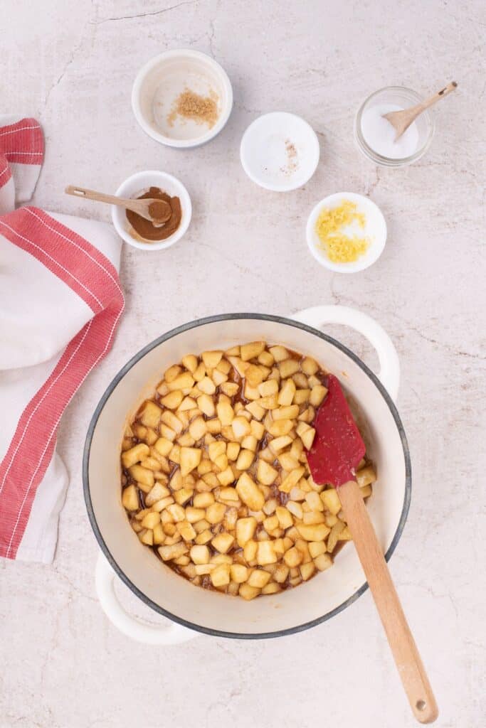 Chopped granny smith apples in a dutch oven with a spatula. Surrounded around small bowls of lemon zest, nutmeg, cinnamon, brown sugar, and salt.