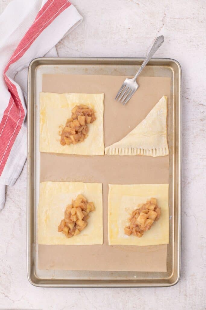 Baking sheet with parchment paper lining it. 4 squares of puff pastry are lined on the parchment with a small amount of apple filling in each center. The puff pastry square at the top right is folded in half on the diagonal with fork pressings on the sides to seal the puff pastry shut.