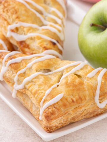 Two completed baked and iced apple turnovers on a white plate with a green granny smith apple in the background.