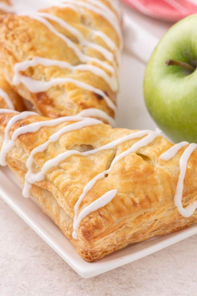 Two completed baked and iced apple turnovers on a white plate with a green granny smith apple in the background.