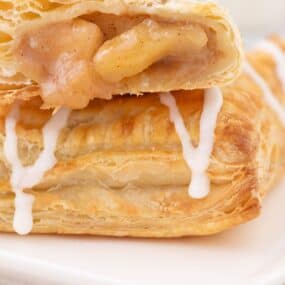 Stacked apple turnovers on a white plate. The top apple turnover is sliced open to show the baked filling inside.