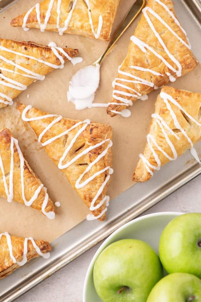 Parchment lined baking sheet with baked and iced apple turnovers. Spoon with icing sets in the center of the turnovers, a bowl of green apples in the bottom corner.