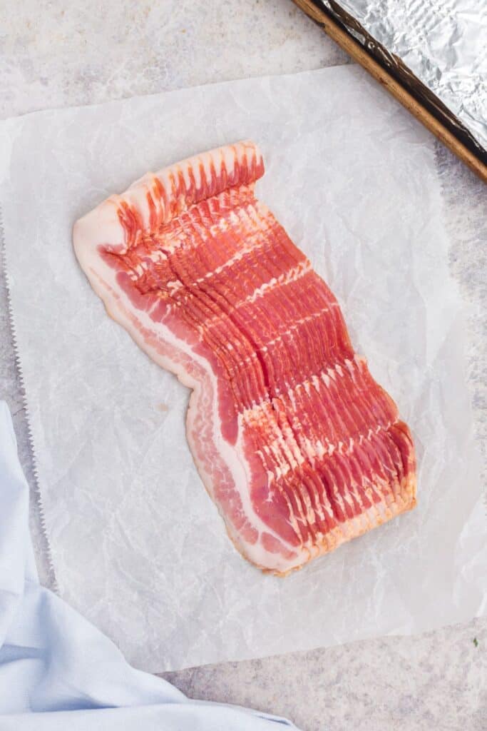 Slab of raw bacon on a sheet of parchment. Surrounded by a towel and a prepped baking sheet covered in aluminum foil.