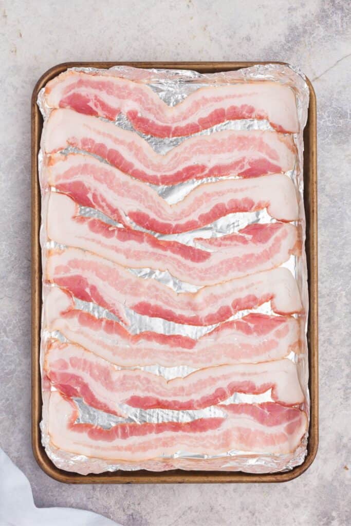 Raw slices of bacon on a baking sheet covered in aluminum foil.