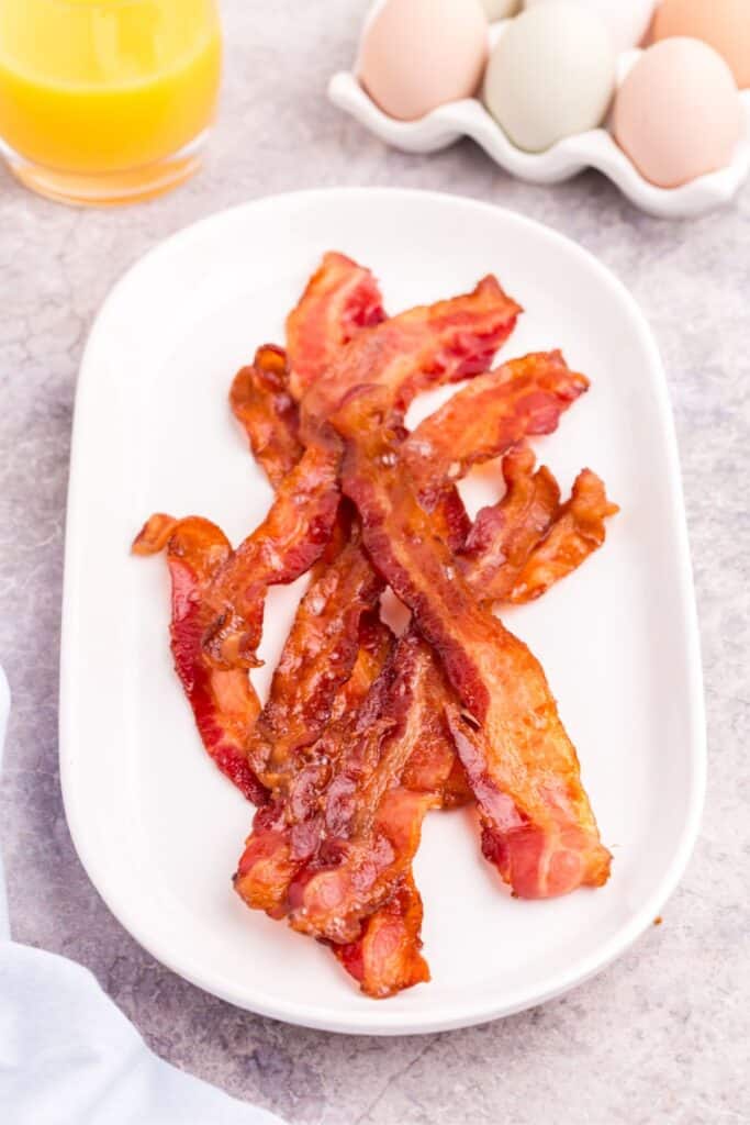 baked bacon on a plate surrounded by a half dozen whole eggs, orange juice, and a napkin