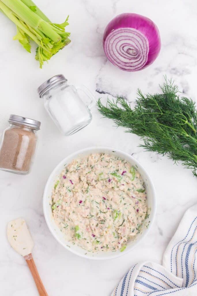 Flatlay of Tuna salad mixture surrounded by celery stalk, half a red onion, fresh dill, salt and pepper.