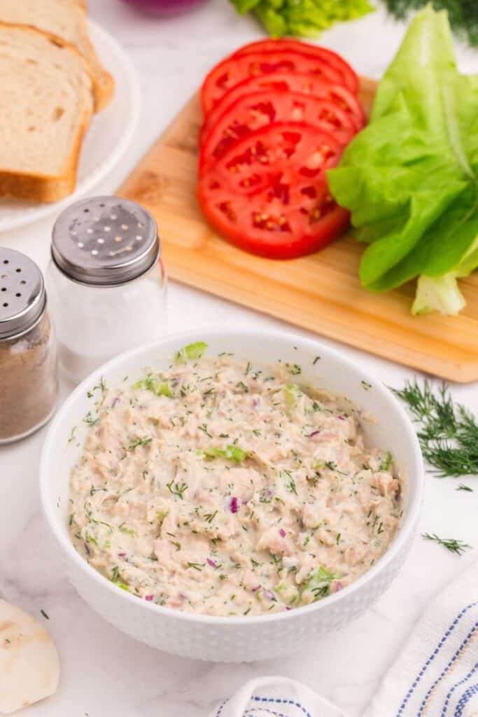 Tuna salad mixture in a bowl. Background includes bread, tomatoes, lettuce, salt, and pepper.