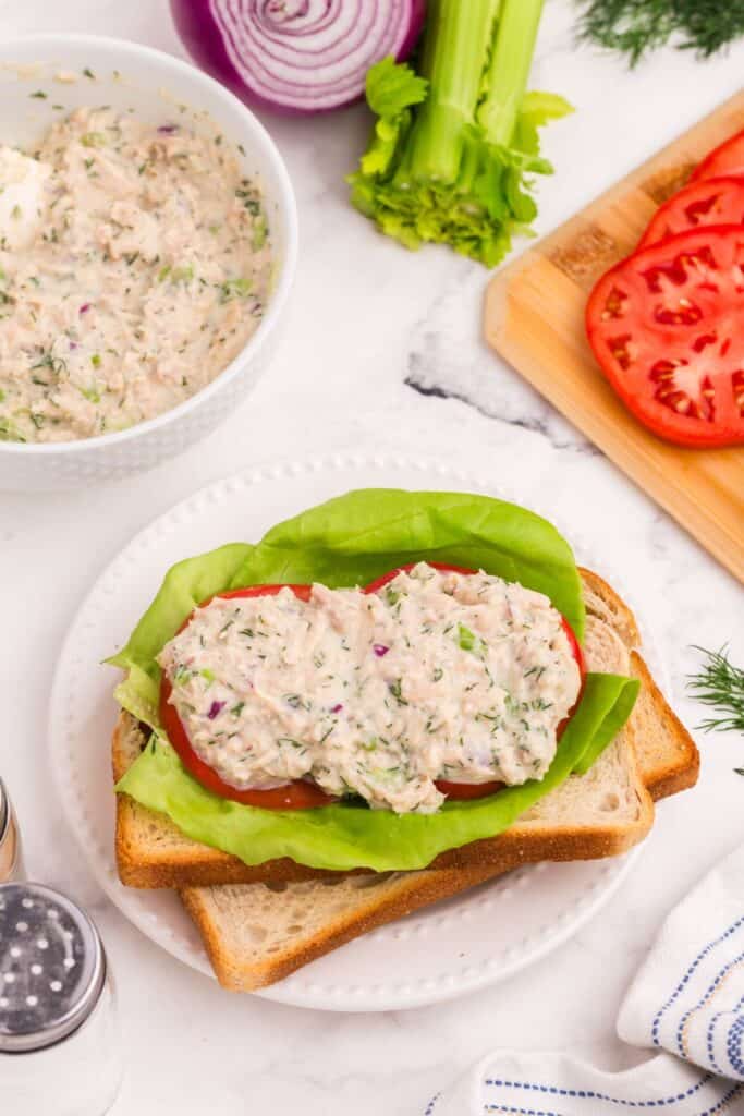 Open faced tuna salad sandwich surrounded by bowl of tuna salad, red onion, celery stalk, and cutting board with sliced tomatoes.