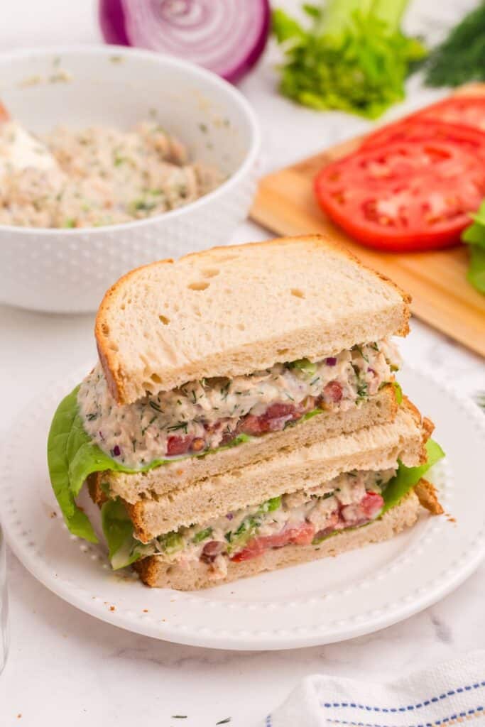 Two halves of a tuna sandwich stacked on one another on a white plate. Bowl of tuna salad in the background with red onion, celery stalk, and cutting board with sliced tomatoes.