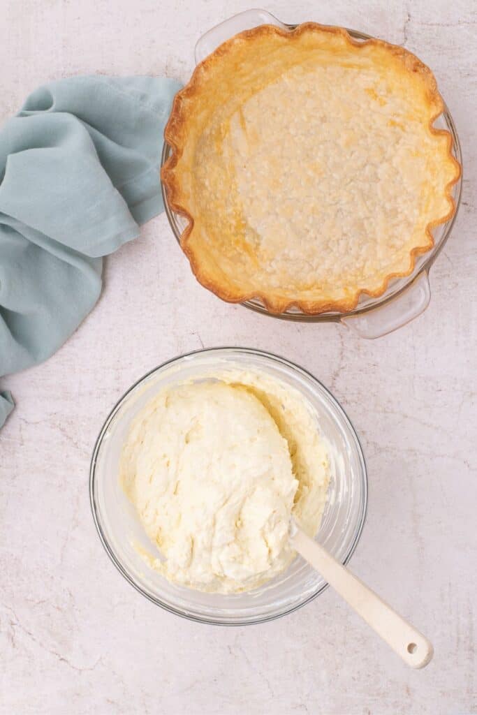 Coconut cream pie mix in a glass bowl with a spatula folded in next to a pre-baked empty pie shell.