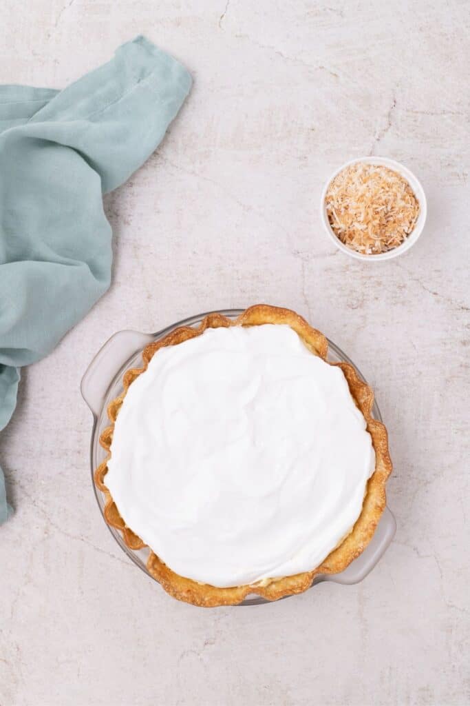 Coconut cream pie mix assembled in the baked pie shell. A bowl of toasted shredded coconut is next to the pie shell.