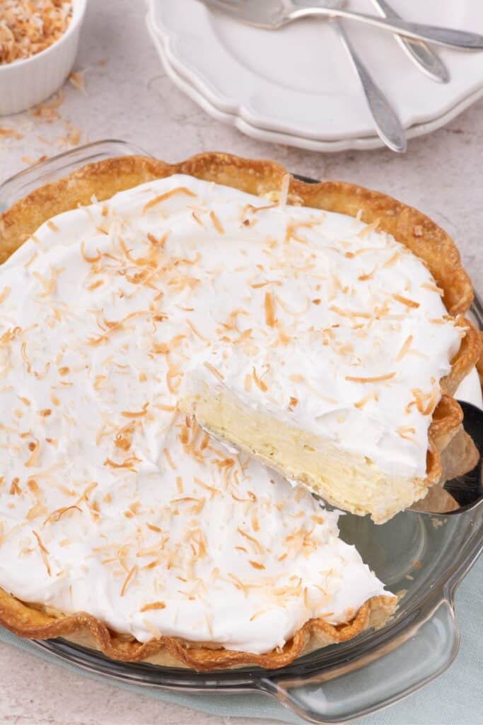 Slice of Coconut Cream Pie lifted above the whole pie. Plates and forks in the background with a bowl of toasted coconut.