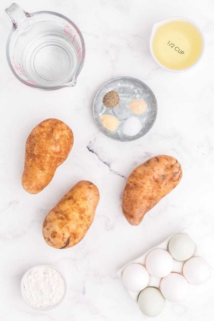 Flatlay for Hash brown recipe. Whole russet potatoes surrounded by half a dozen whole eggs, flour, a tray of spices, a measuring cup of water, and a small bowl of oil.