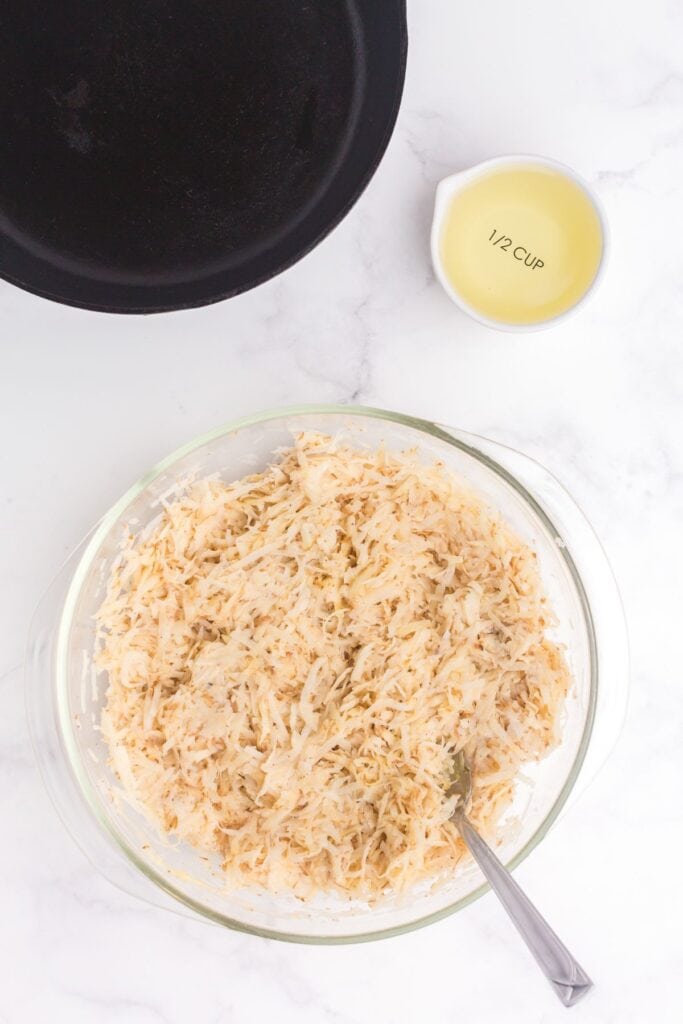 Glass bowl with Hash brown mixture in it. Cast iron pan and small bowl of oil above the hashbrown mixture.