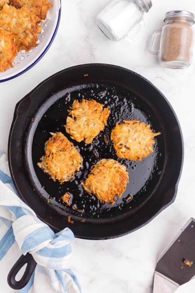 Four small hash brown patties frying in a well oiled cast iron pan.