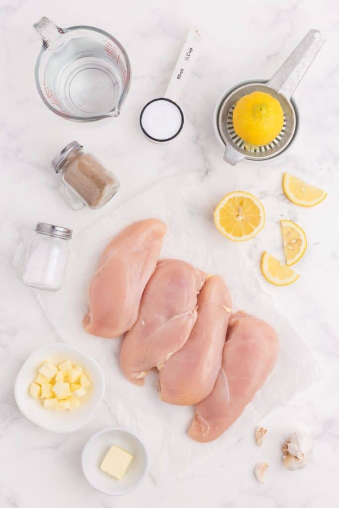 Flatlay of raw chicken surrounded by lemons, salt, pepper, butter, and garlic