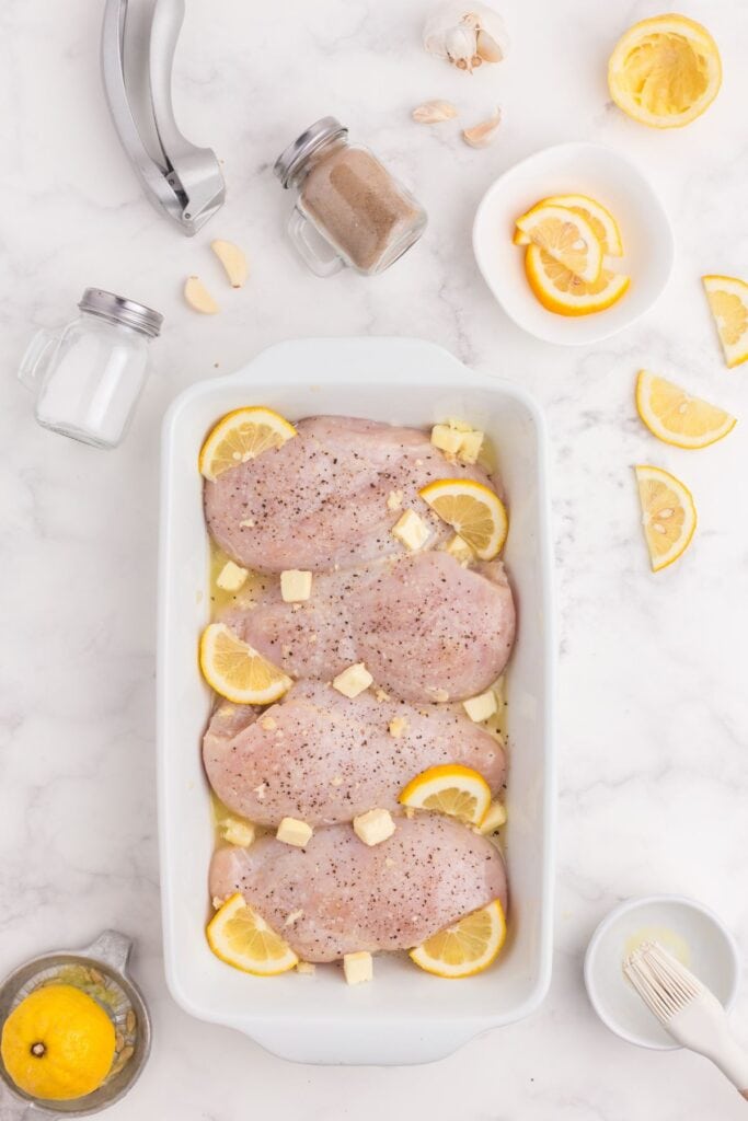 Raw chicken arranged in a baking dish surrounded by lemon wedges, salt, pepper, garlic, garlic press, and a basting brush