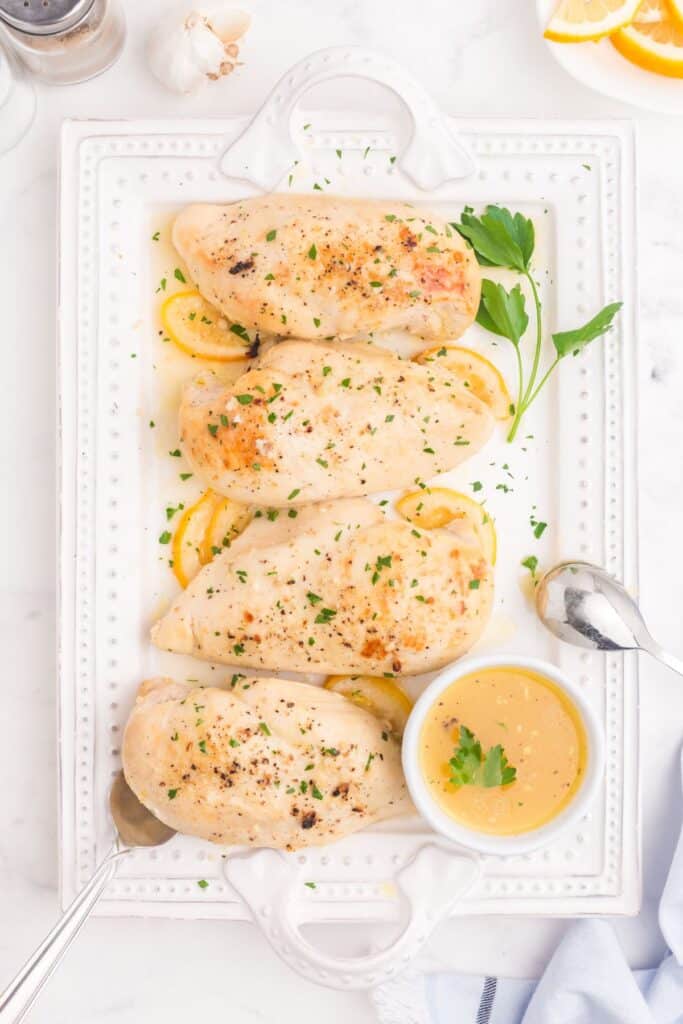 Pulled back view of cooked lemon chicken on a serving tray with lemon and parsley garnishes. Served with a small bowl of lemon butter on the side.