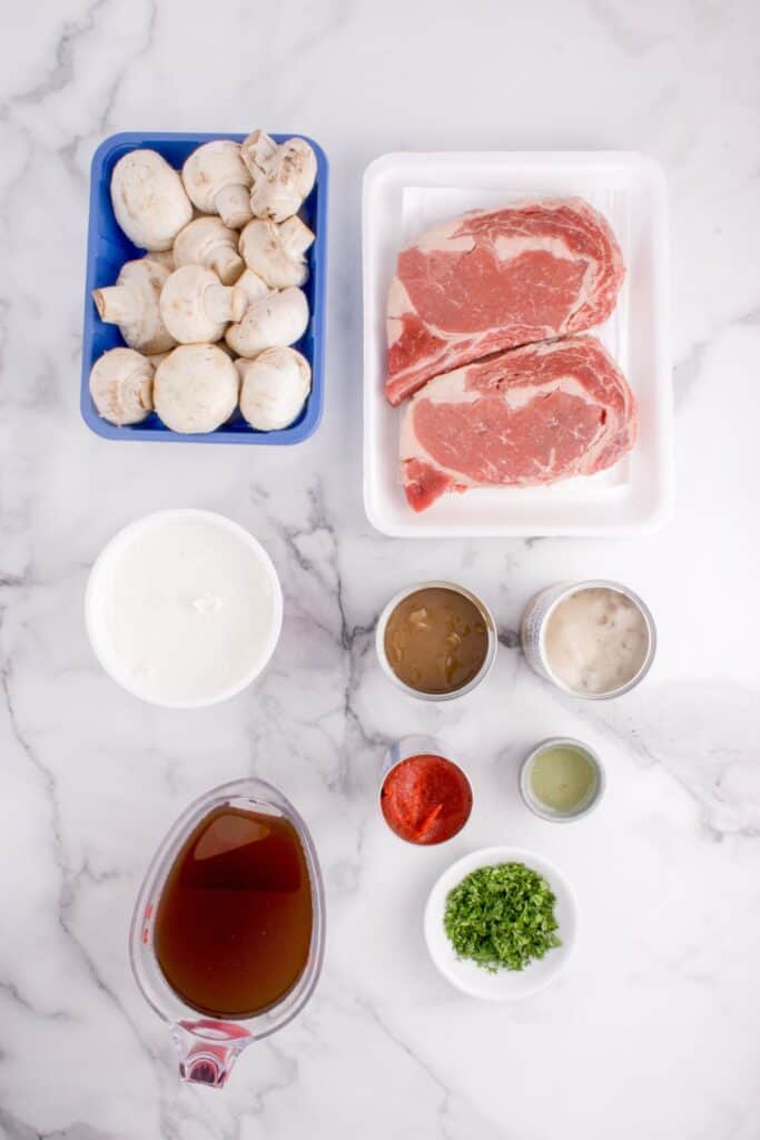 Flatlay of Beef stroganoff ingredients. Boneless ribeye steaks, white button mushrooms, can of cream of mushroom soup, can of brown gravy with onions, sour cream, tomato paste, beef broth, parsley and olive oil.