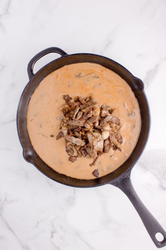 Progress image of Beef stroganoff recipe, with thinly sliced ribeye steak added in a pile in the center of the cast iron pan.