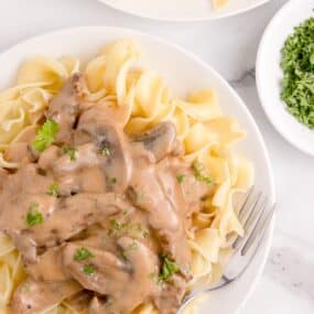 Beef stroganoff served over a bed of egg noodles on a white plate with a fork. Parsley garnish on top.