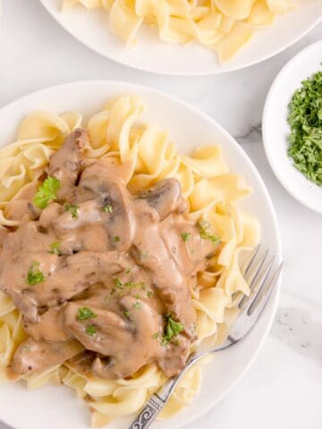 Beef stroganoff served over a bed of egg noodles on a white plate with a fork. Parsley garnish on top.